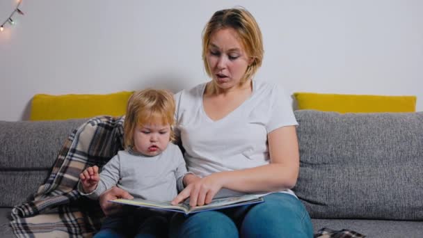 若い母親がソファに座っている間に子供に本を読んでいる 就学前教育 スローモーション — ストック動画
