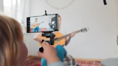 A young mother playing the guitar and records a live video broadcast on a smartphone. Child plays with smartphone. The concept of video streaming and e-learning.