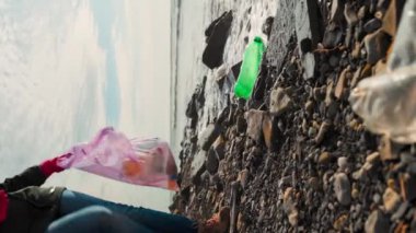 Kadın bir gönüllü çöp torbasına plastik bir şişe koydu. Dikey oryantasyon. Düşük açı. Dünya Çevre Günü kavramı ve vahşi kıyıları temizlemek