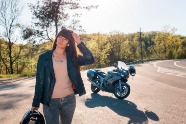 Dünya Motosiklet Günü konsepti. Deri ceketli, elinde motosiklet kaskı tutan ve güneş gözlüğü takan genç bir kadın. Arka planda bir motosiklet ve yol.