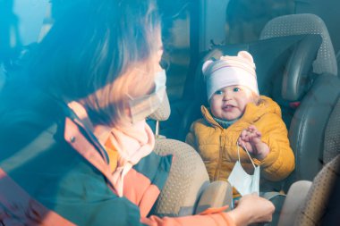 Koruyucu maskeli bir annenin portresi, arabasında oturuyor ve çocuğuna maske takıyor çocuk oto koltuğunda arka koltukta oturuyor. Ön camdan bak..