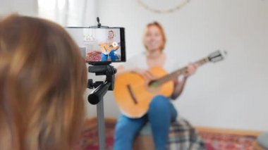 A little daughter helps her mother record a live online broadcast of playing the guitar. The child adjusts the smartphone standing on a tripod. Close-up from the child's shoulder