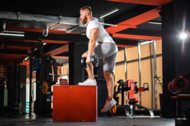 Young bearded athletic man training in the gym jumping at cube with dumbbells. The concept of fitness and work out.