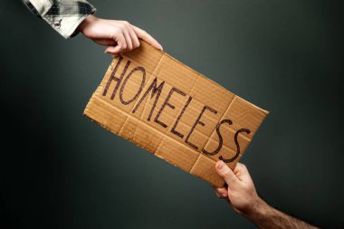 A woman and a man hold a cardboard sign with the inscription HOMELESS. Dark background. Hands close-up. The concept of helping vagabonds and panhandle.
