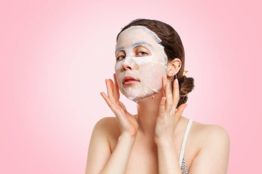 Portrait of a young beautiful Caucasian woman posing with a fabric moisturizing mask on her face. Pink background. Copy space.The concept of beauty, hydration and self-care.