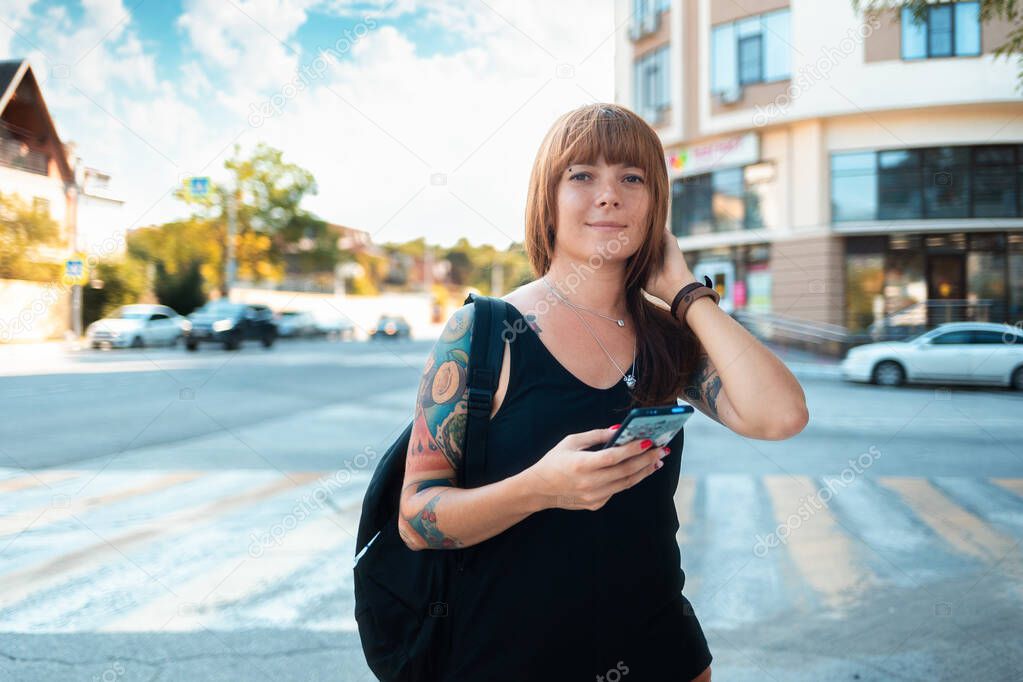 Portrait of a young smiling woman with a sleeve tattoo standing on the street and holding a smartphone. Street in the background. The concept of freedom and travel.
