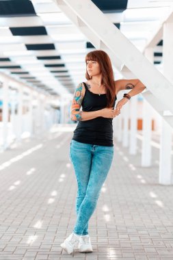 A young beautiful woman with tattoos on her arm, posing, standing near a white staircase. In the background is an architectural perspective. Vertical. Outdoor.