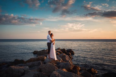 Wedding Day. Newlyweds embrace and pose standing on the coastal rocks. In the background, the sunset and the ocean and the sky are in the clouds.