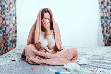 The concept of colds. A woman sits on the bed covered with a blanket, and blows her nose in a handkerchief. Next to her are pills, a thermometer and handkerchiefs. Window in the background. Copy.
