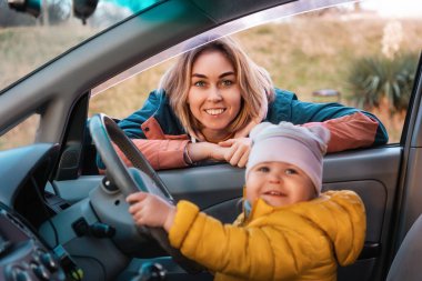 Portrait of a young smiling Caucasian mother looking through a car window. A happy kid is sitting in a car seat and holding the steering wheel with his hands. Side view. The concept of driving training.