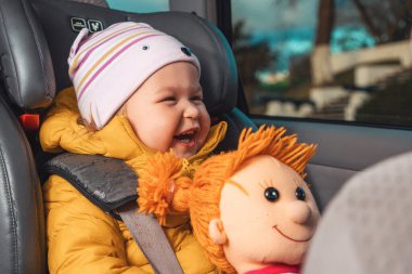 Portrait of happy little smiling caucasian child is sitting strapped in a child seat with a toy in his hands. View inside a car.