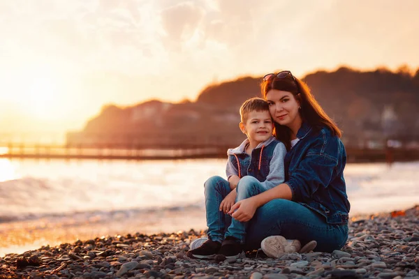 一位年轻的母亲和她的儿子坐在一起 在海滩上拥抱 背景是大海和日落 复制空间 幸福童年的概念 — 图库照片