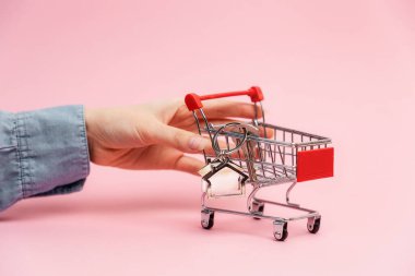 Insurance of real estate. Female hand push shopping mini cart with key and house keychain in it. Pink background. Concept of leasing of real estate and mortgage.