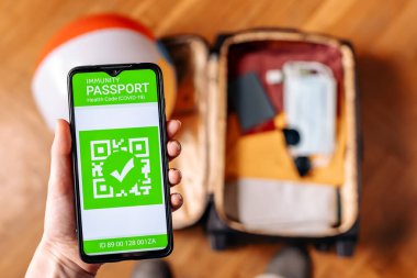 The concept of new normal. Close up view of a male hand holds a smartphone with an immune digital health passport. Top view. A suitcase with clothes and protective masks on the blurred background.