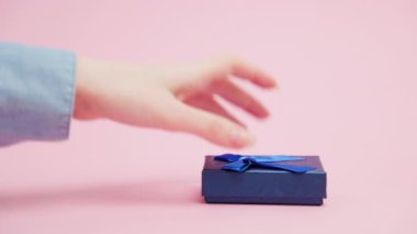 Close-up of a woman's hand in shirt opens gift box and shows a new keys with a metal keychain house. Pink background. The concept of mortgage and buy real estate.