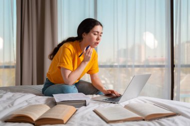 Pretty woman is sitting on a bed and working on a laptop. Books are lying next to each other. The concept of online courses and remote work.