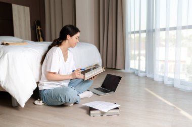 A young woman sits on the floor and studies from a book. There are papers and a laptop on the floor. The concept of internet education and online courses.