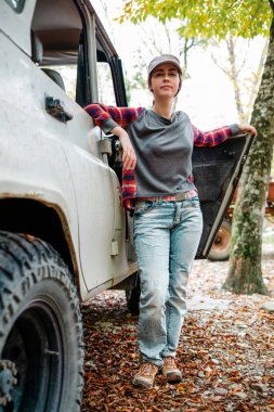 A young Caucasian woman poses next to a jeep. Trees in the background. Vertical. Concept of car travel and tourism.