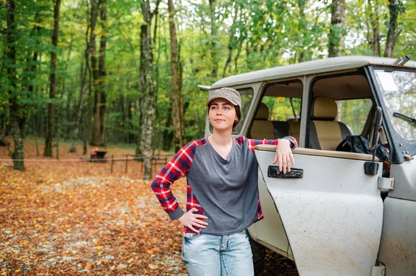 A young Caucasian woman poses next to a jeep and looks away. In the background, trees and forest. Concept of car travel and tourism.