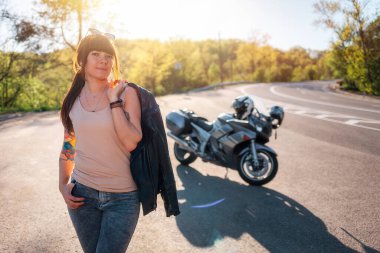 Deri ceketli şık dövmeli bir kız yolun kenarındaki motosikletin arka planında poz veriyor. Motosiklet yerel seyahat konsepti ve Dünya Motosiklet Günü.