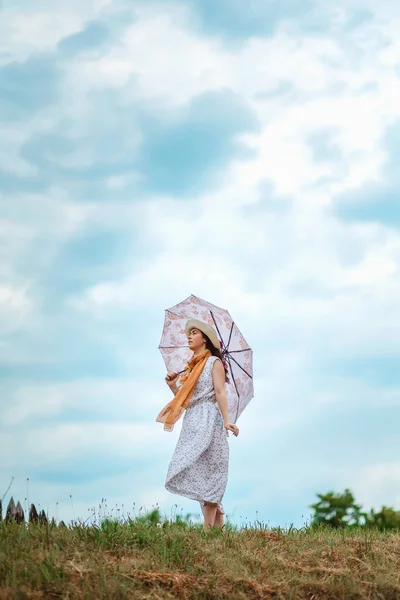 一个戴着草帽 穿着衣服 手里拿着雨伞的年轻女子站在山上 望着多云的天空 复制空间 心理学的概念 — 图库照片
