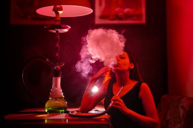 A sexy elegant woman smokes a hookah and lets out a cloud of smoke. Red tint.