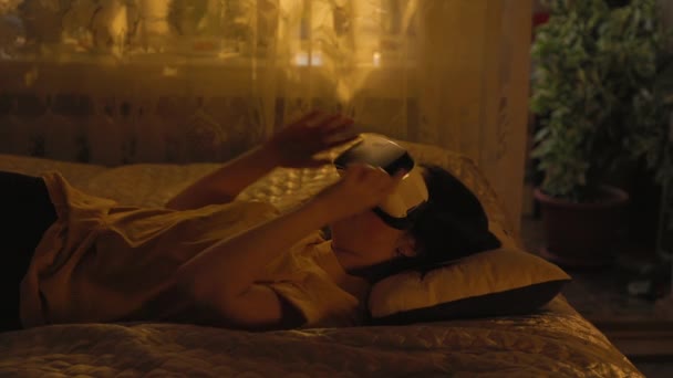 一个年轻的高加索女人躺在床上 戴着一副虚拟现实眼镜 在家里玩游戏 — 图库视频影像