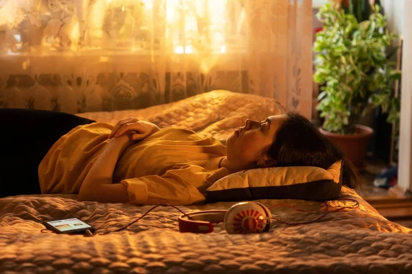 眠い若い女性をリラックス枕 携帯電話 ヘッドフォンのベッドの上に横たわっている近くにあります 家で冷静に 窓からの夕日の光 — ストック写真