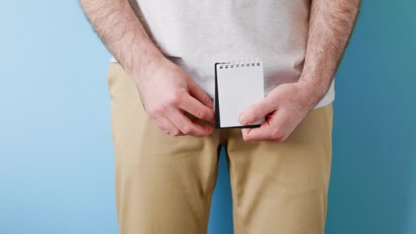 男性生殖系统疾病 一个穿着米色牛仔裤的男人手里拿着一本笔记本 上面有两只手的问号 靠近点蓝色背景 人工授精和性无能的概念 — 图库视频影像