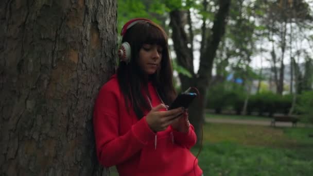 一位年轻漂亮的女士站在公园的一棵树旁 她用智能手机 用耳机听音乐 潘妮开枪了公园内的康乐 — 图库视频影像