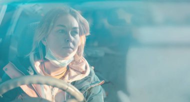 Arabanın direksiyonunda oturan koruyucu maske takan genç bir kadının portresi. Ön camdan dışarı bak. Uzayı kopyala Yeni normal kavram.