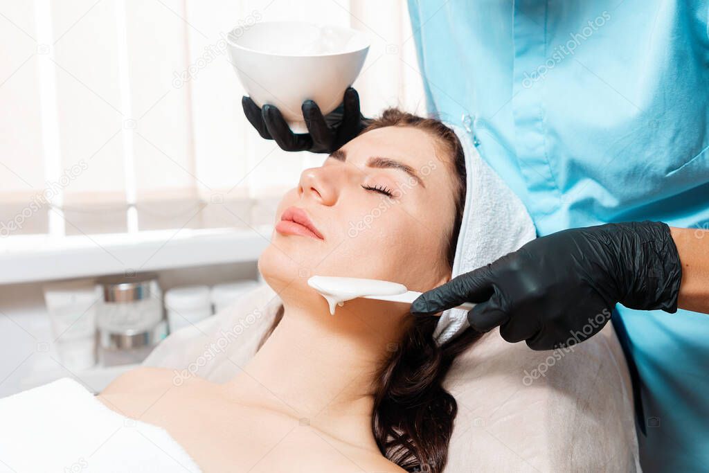 Beauty procedure. Cosmetologist in black latex gloves applying with spatula algae mask on the woman's face. Concept of professional cosmetology and skin care.