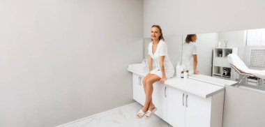 Genç, gülümseyen bir kozmetikçi kadın bir tıp ofisinde masanın üzerinde oturuyor. Uzayı kopyala Profesyonel kozmetoloji kavramı.