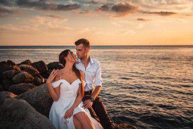 Düğün günü. Yeni evliler sahildeki kayalıklarda otururken kucaklaşıp poz verirler. Arka planda, günbatımı, okyanus ve gökyüzü bulutlarda. Sevgililer Günü.