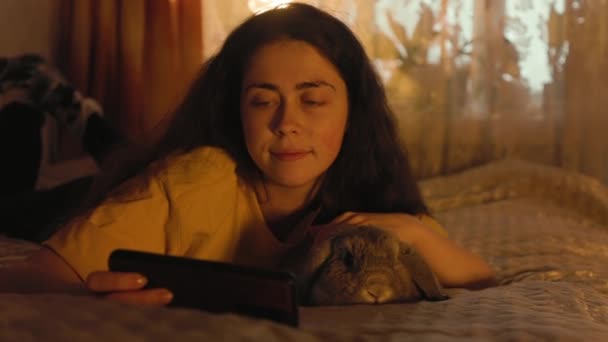 一个微笑的女人躺在床上摸着一只失聪的兔子 看手机的画像 宠物狗照顾 家庭娱乐 — 图库视频影像