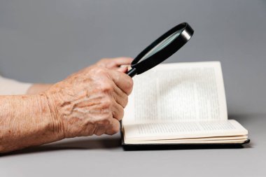 Yaşlı bir kadının buruşuk elleri büyüteçle kitap okuyor. Gri arka plan. Araştırma ve eğitim kavramı.