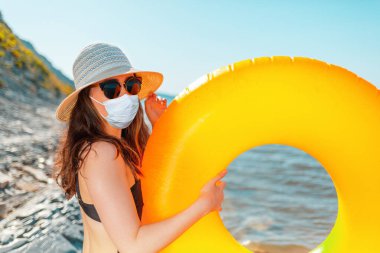 Mayo, şapka ve tıbbi maske takan bir kadın denizin arka planında turuncu şişme bir daire tutuyor. Tatilin boyunca virüs koruması kavramı.
