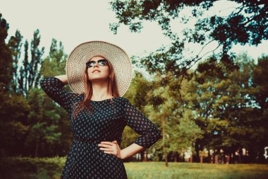 Beyaz şapkalı ve mavi elbiseli kız, açıklıkta dikiliyor, ağaçların arka planında..