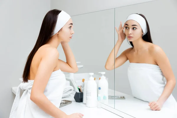 若い美しい女性がバスルームに立って鏡の中で彼女の顔を見ています スキンケアと若返りの概念 — ストック写真