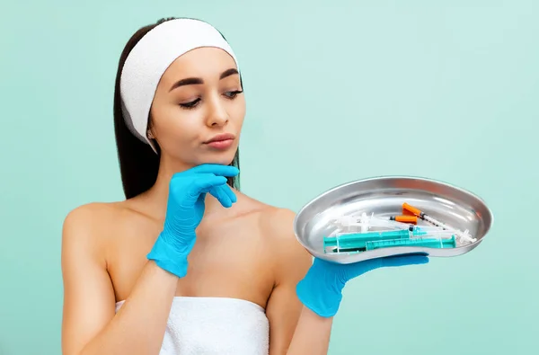 Portrait of a thoughtful young pretty woman in medical gloves choosing syringes from medical kidney dish. Light turquoise background. Concept of plastic surgery.