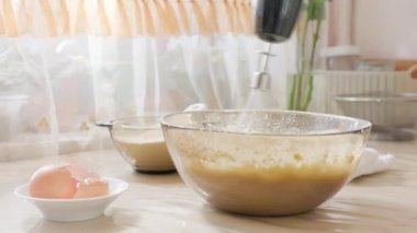 Bir dişinin elinin yakınında çırpılmış hamur ve cam bir kase içinde mikser var. Yanındaki masada beyaz bir havlu, bir mikser ve yumurta kabuğu var. Yemek pişirme konforu ev yapımı gıda konsepti