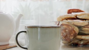 Sıcak çayla seramik bardağı kapat. Arka planda porselen beyaz bir çaydanlık ve bir kase krep var. Ev çay partisi.