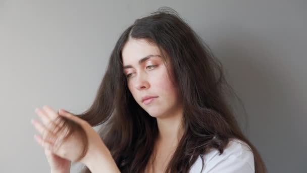 Retrato de uma jovem mulher branca bonita olhando para as pontas secas de seu cabelo. Fundo cinzento. O conceito de cuidado do cabelo. — Vídeo de Stock