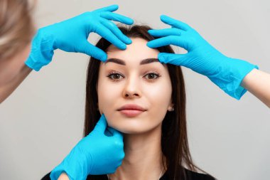 Lastik eldivenli iki kozmetik uzmanı bir kadının yüzünü inceliyor. Genç ve güzel bir kadının portresi. Profesyonel cilt bakımı ve yüz tazeleme kavramı.