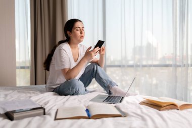 Genç, beyaz bir kadın bir yatakta oturuyor ve akıllı telefon kullanıyor. Uzayı kopyala Çevrimiçi eğitim ve sosyal ağlara bağımlılık kavramı.