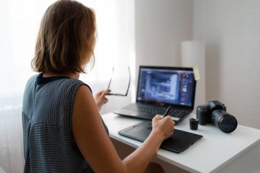 İş ve yaratıcılık. Gözlüklü genç bir sarışın rötuş yapıyor, masada bir kamera var. Arka plan.