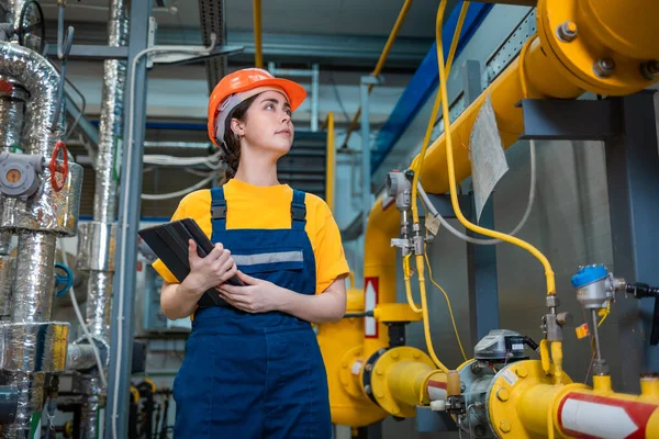 Production inspection. A young engineer in a uniform and a protective helmet, holding a tablet in his hands and conducting an inspection of the equipment.