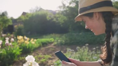 Hasır şapkalı gülümseyen genç bir kadın akıllı telefonunda çiçek açan irislerin fotoğraflarını çekiyor. Açık havada. Yaz bahçesi.