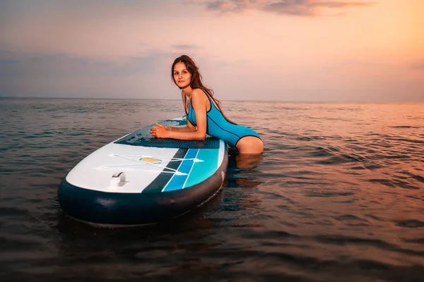 水着姿の若いスリムな女性が板の上に腰を下ろしている 背景に夕日 コピースペース ウォータースポーツと夏のアクティビティの概念 — ストック写真