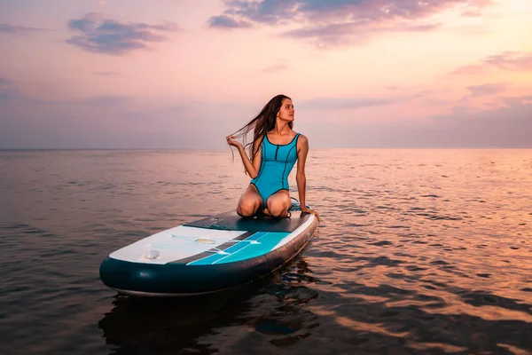 穿着泳衣的年轻貌美的女人坐在一个冲浪板上 美丽的落日和背景中的海洋 — 图库照片
